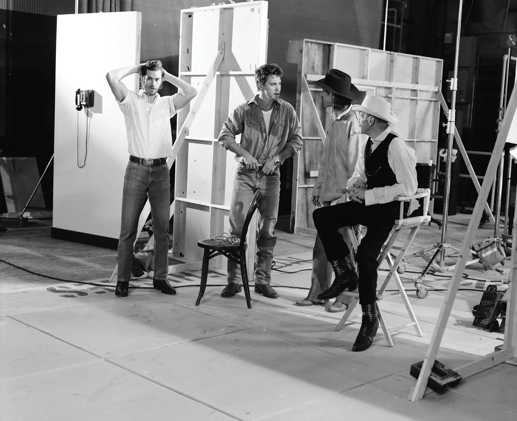 Behind-the-scenes B&W shot of three adult men on set, one seated in a director's chair, all wearing casual western-style clothing.