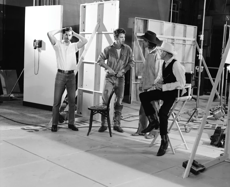 Behind-the-scenes B&W shot of three adult men on set, one seated in a director's chair, all wearing casual western-style clothing.