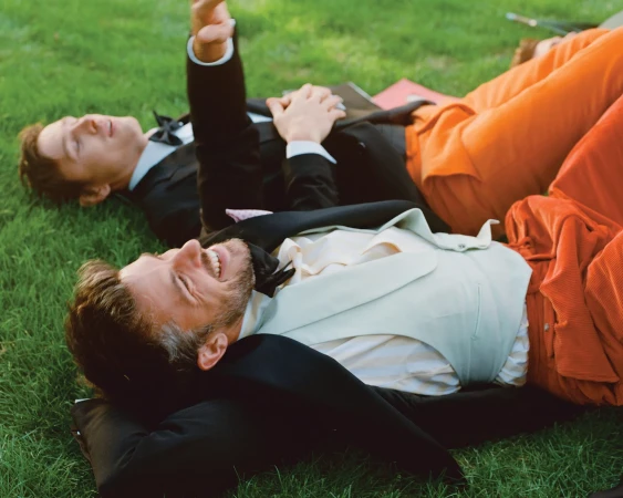 Two adult men in formal wear lounge on the green grass, one in a classic black tuxedo and the other in an orange suit, smiling in daylight.