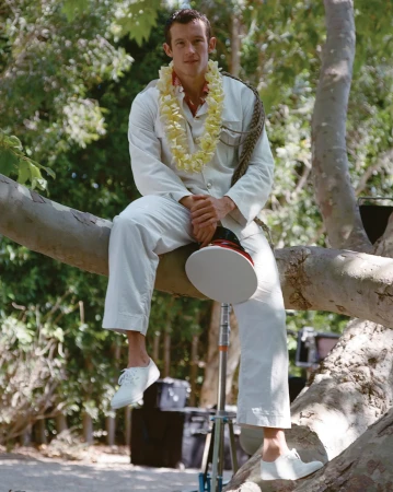 Man in white suit sits on tree branch wearing a lei in lush outdoor setting, holding a film reflector, trees in background.