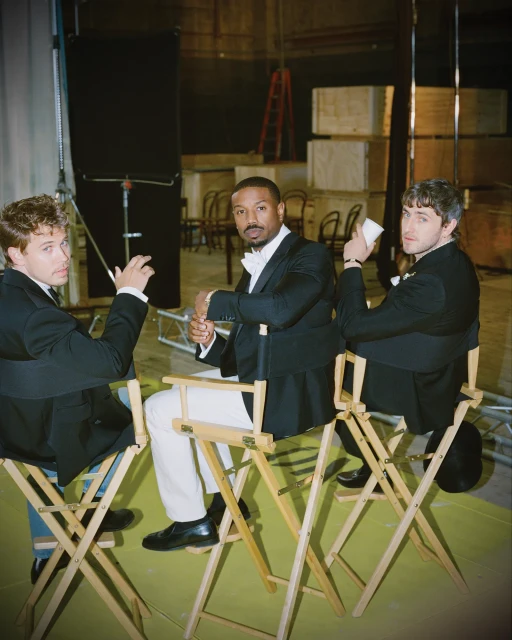 Three men in formal wear pose for a photo in an industrial setting, sitting on director's chairs, with metal staging in the background.