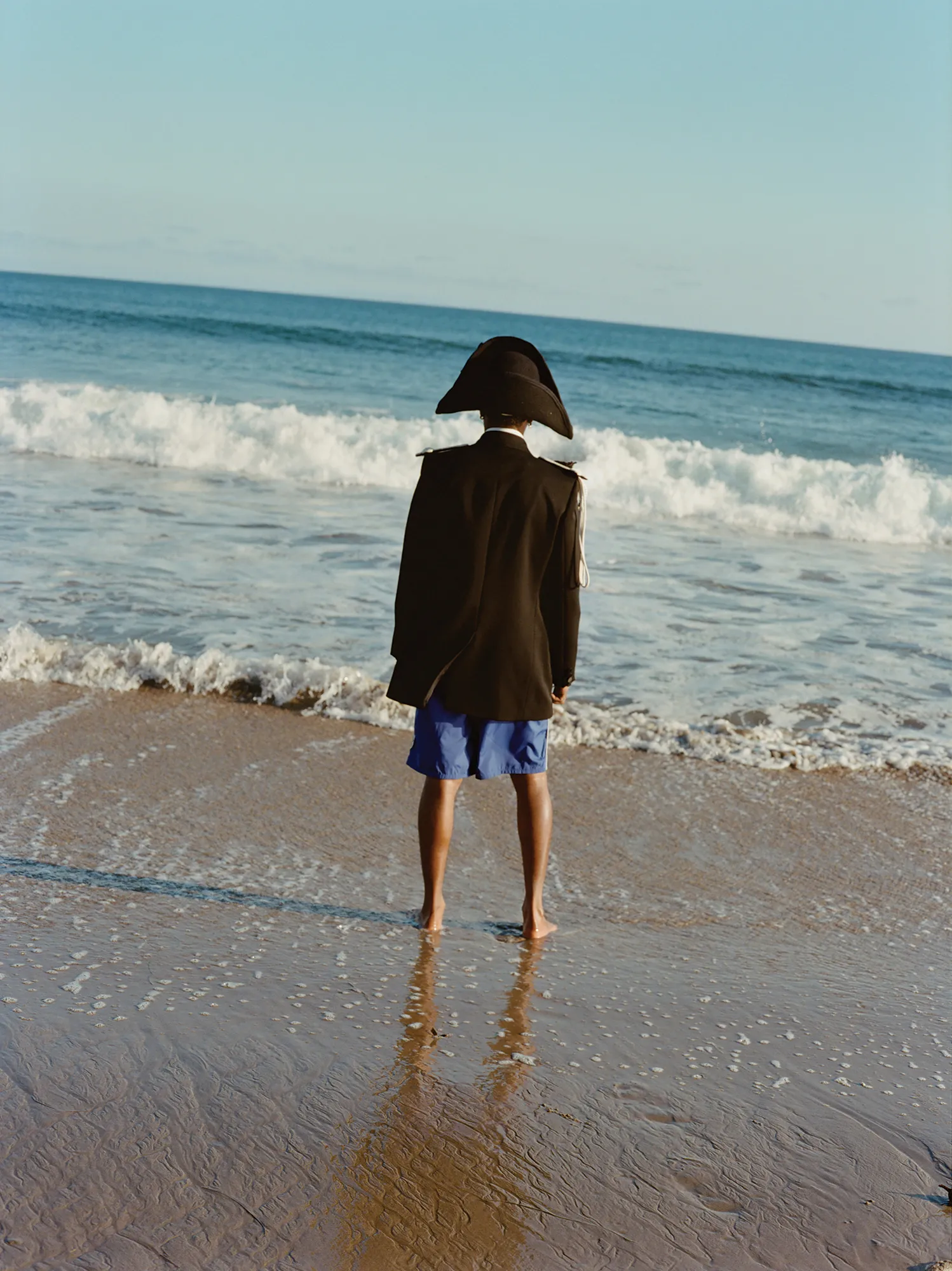 Person in period hat and jacket on a beach, facing ocean waves, reflecting in wet sand. Sunny day, seascape, water, hat, and historic style.