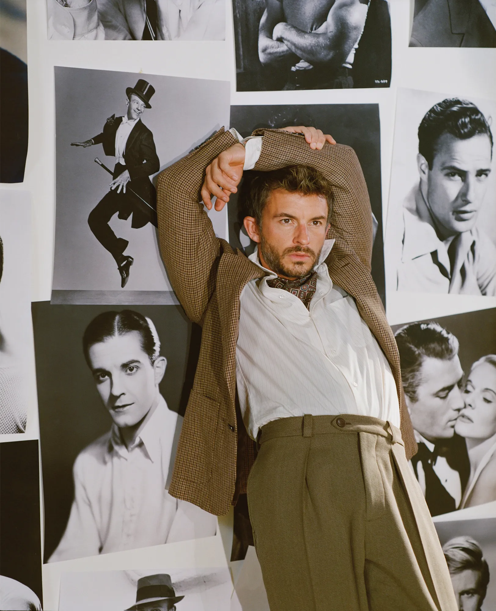 Adult man in jacket and slacks poses with arms raised against wall covered in black-and-white celebrity photographs in neutral light.
