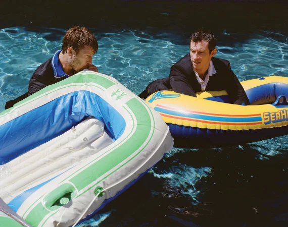 Two adult men in suits sit in inflatable boats floating on water. One man is inflating a boat, while the other looks off to the side.