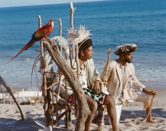 Two men in pirate-like costumes sit on a driftwood structure on a beach next to a scarlet macaw perched on a branch.
