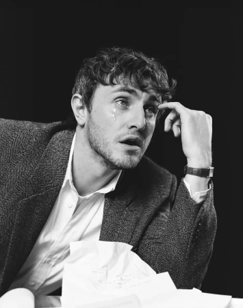 Black and white portrait of a crying man in a blazer and dress shirt, seated at a table with crumpled papers, against a dark background.