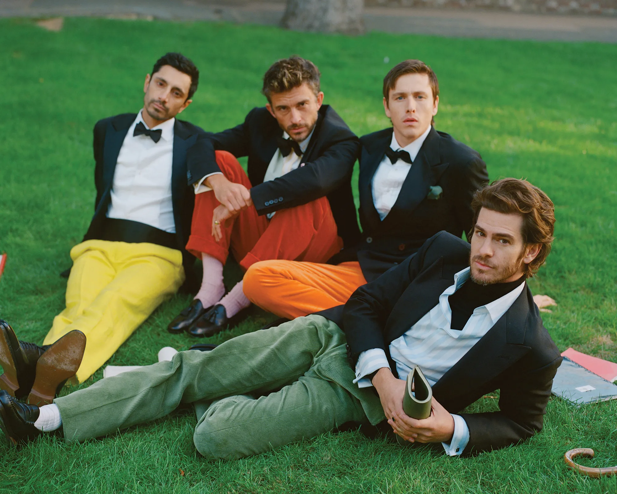 Four men in formal wear sit in grass on a sunny day, exhibiting diverse fashion choices with colored pants and accessories.