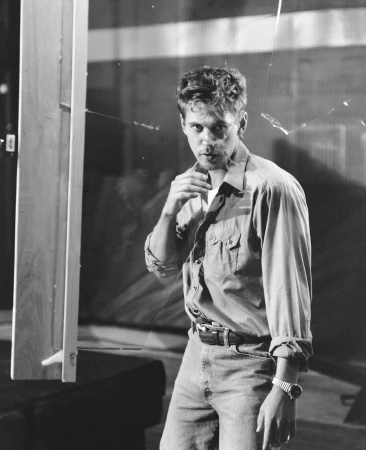 Black and white portrait of a man in shirt and jeans holding his hand up to his throat, next to glass and furniture.