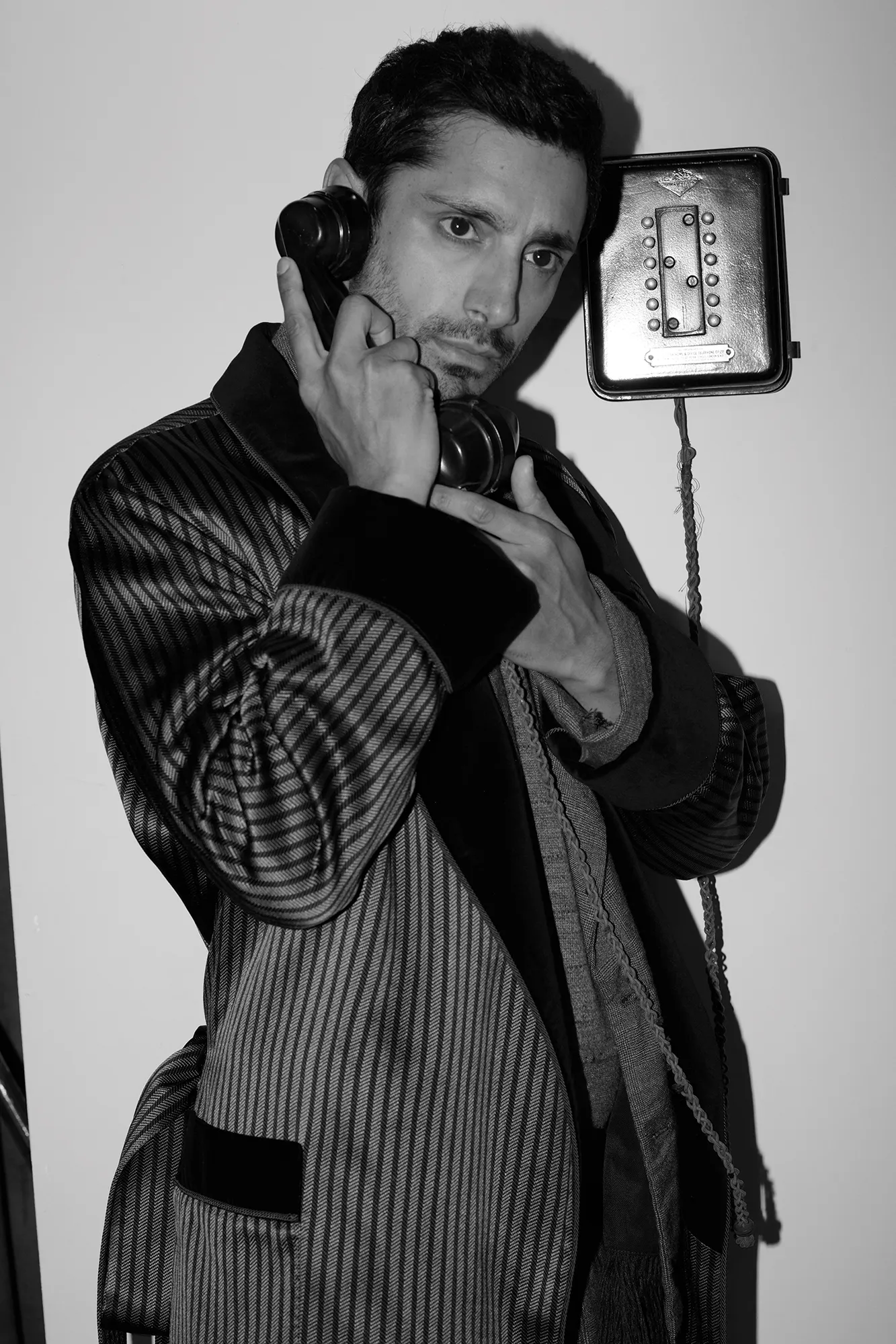 Black and white portrait of an adult male indoors, wearing a robe and holding a vintage telephone receiver to his ear.