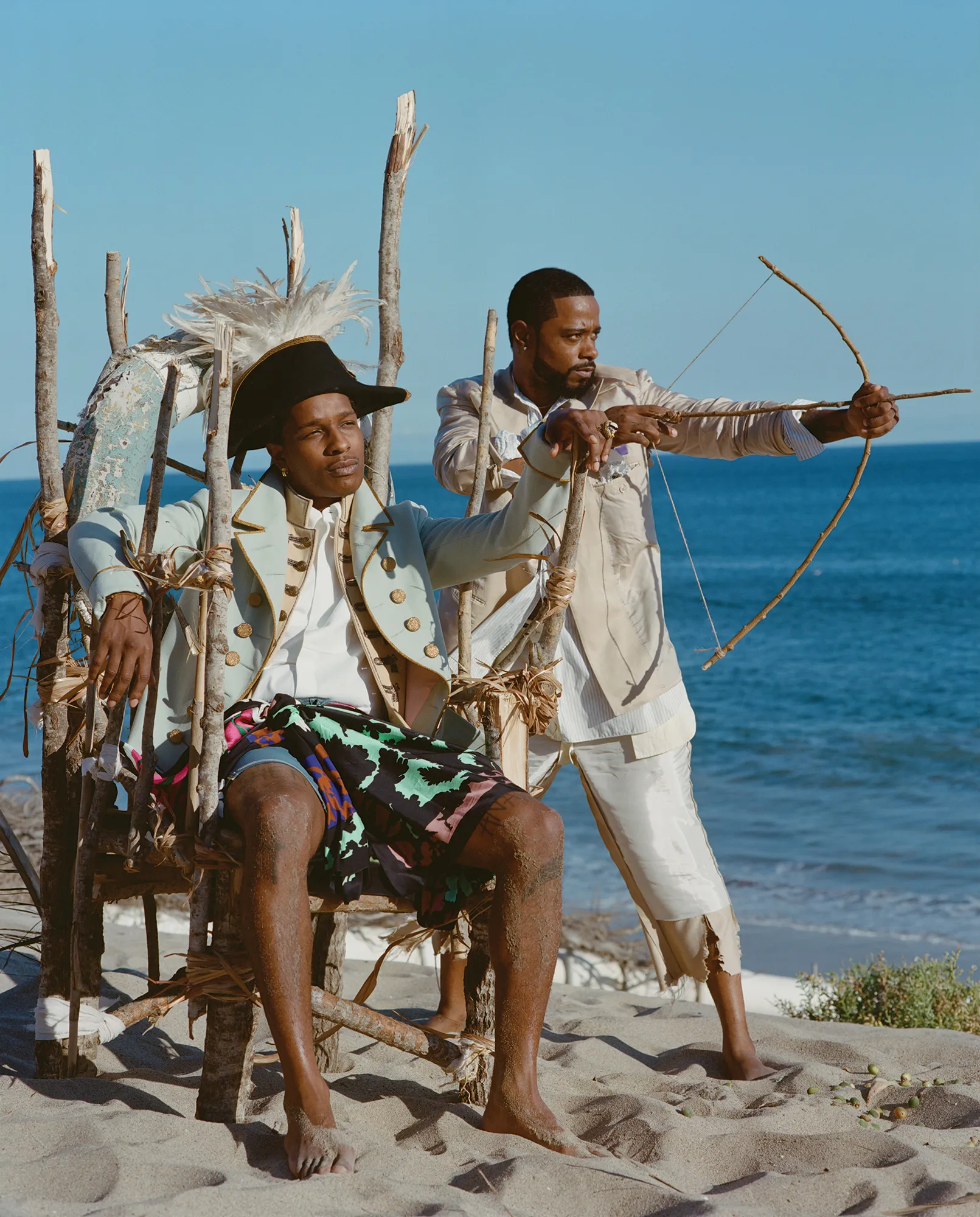 Two men in period costumes posing on a sandy beach. One sits in a wooden chair while the other aims a bow and arrow toward the ocean.