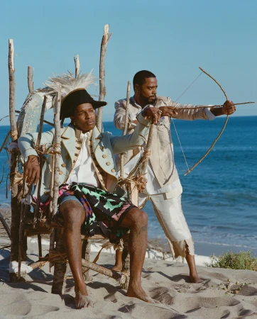 Two men in period costumes posing on a sandy beach. One sits in a wooden chair while the other aims a bow and arrow toward the ocean.