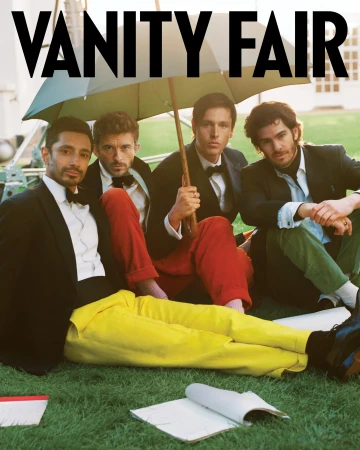 Four men in suits and bow ties are sitting on grass under an umbrella. Red and yellow pants add a touch of color. Vanity Fair logo visible.