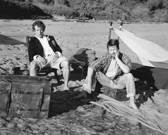 Two men sit on a beach near a makeshift shelter. One man wears a suit jacket, while the other is dressed casually in a plaid shirt.