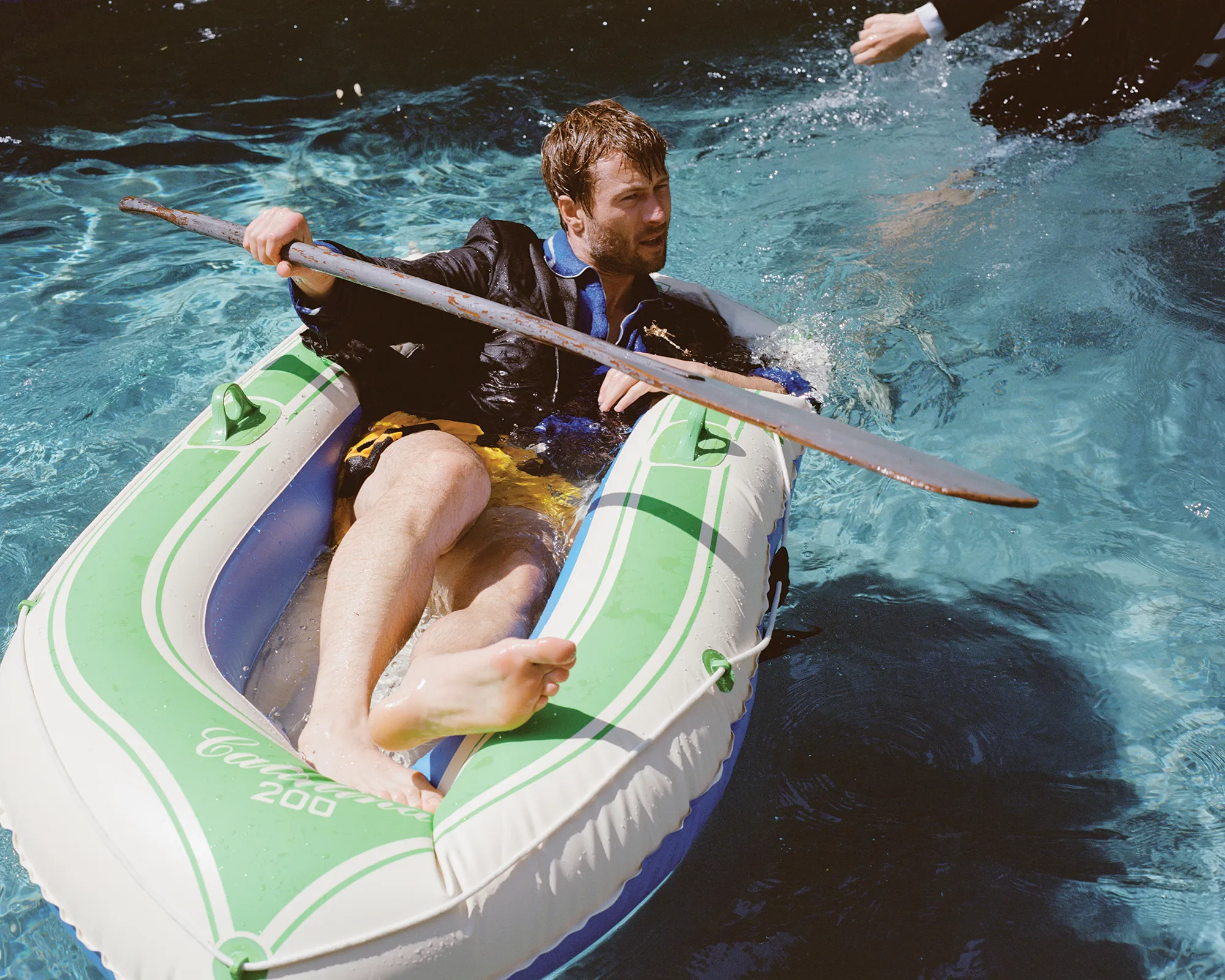 Man in a suit rowing an inflatable boat in a pool. The light is bright and the scene suggests a humorous or unexpected situation.