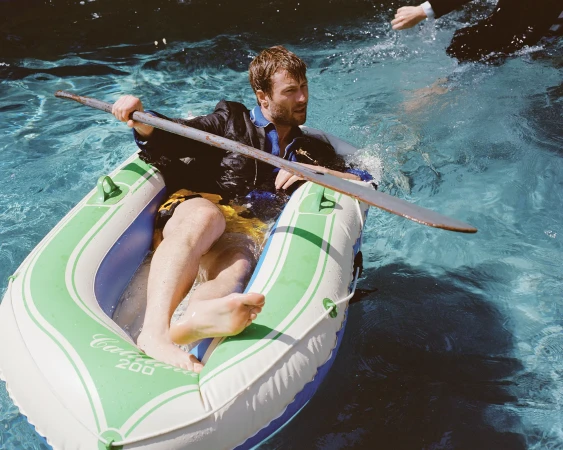 Man in a suit rowing an inflatable boat in a pool. The light is bright and the scene suggests a humorous or unexpected situation.