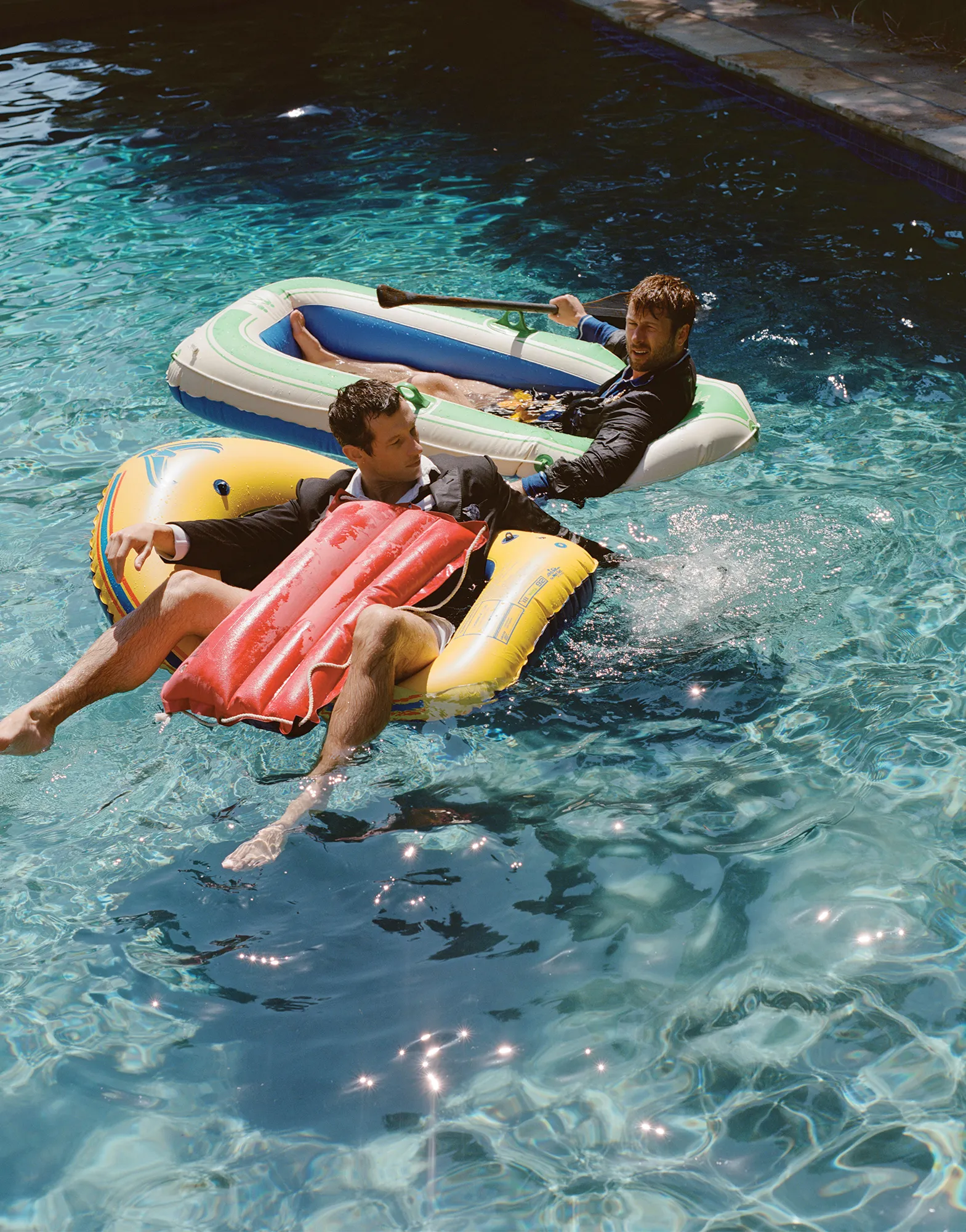 Two men in suits float in a pool on inflatable rafts under sunny lighting, creating a playful and unexpected juxtaposition of formal wear.
