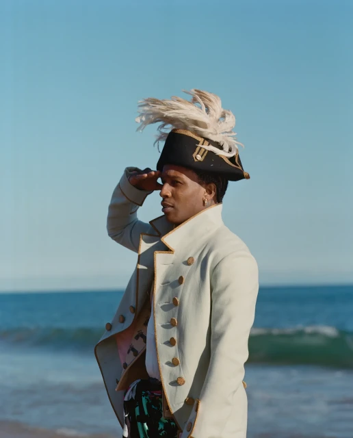 African-American man in a historical military costume poses outdoors at the beach against the ocean and blue sky, saluting.