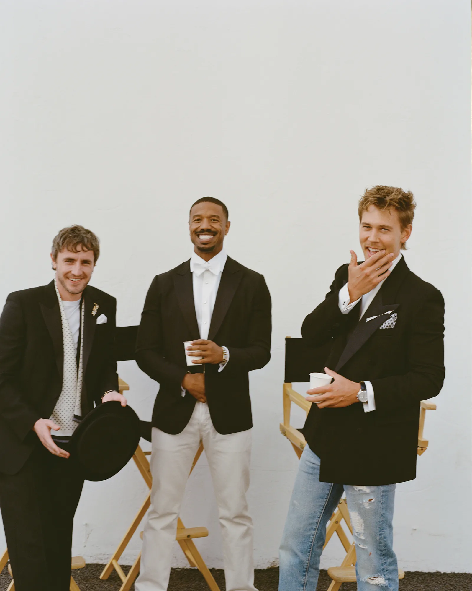 Three adult men in formal wear pose outdoors with director's chairs against a white wall, smiling and holding cups.
