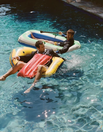 Two men wearing blazers and shorts lounge in colourful inflatable boats on a sunlit outdoor swimming pool.