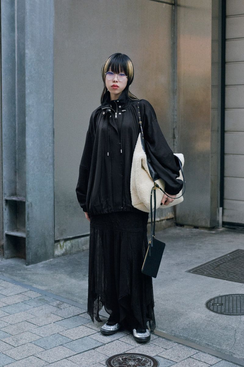 A stylish adult female stands on a city sidewalk in a black jacket and long sheer skirt, accessorized with glasses and a handbag.