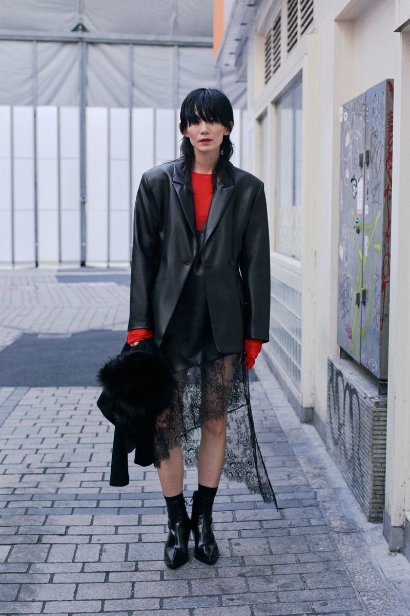Stylish woman with short dark hair wearing a black blazer and lace skirt, posing on a brick walkway in an urban environment.