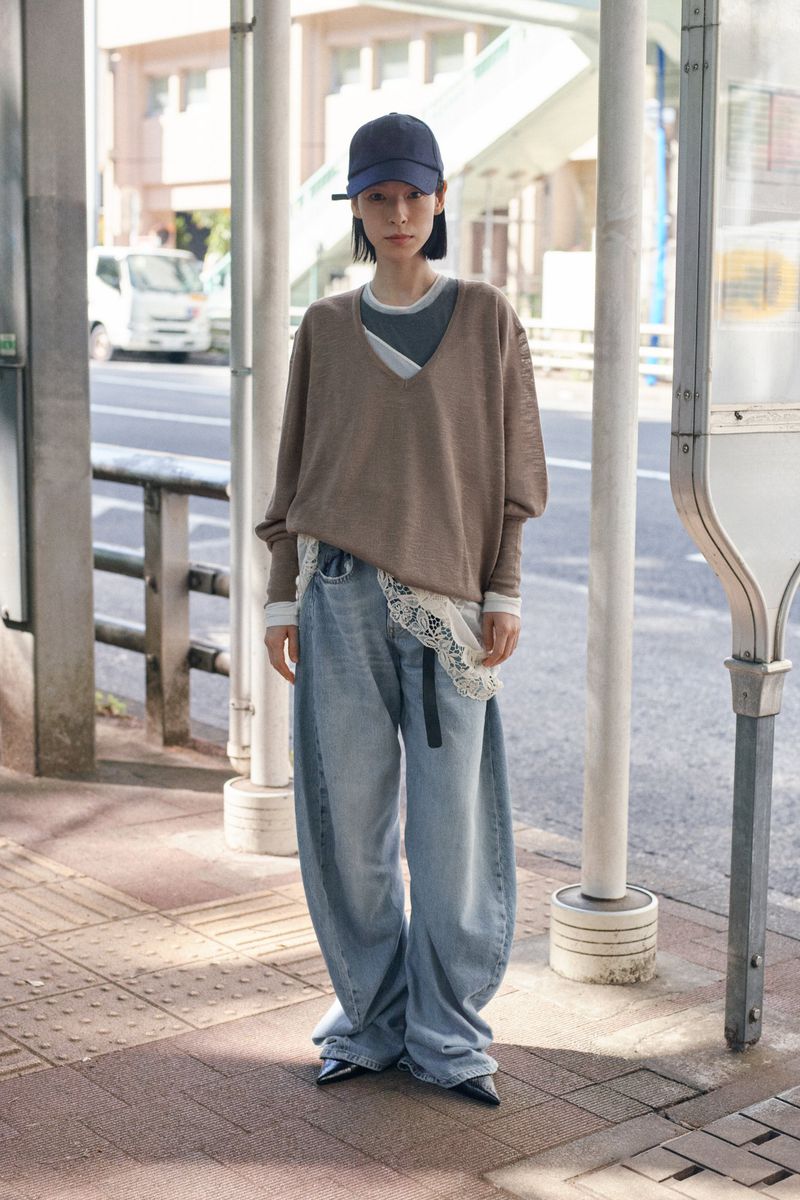 Woman in oversized jeans and a beige sweater stands at a bus stop, showcasing casual fashion in an urban setting on a sunny day.
