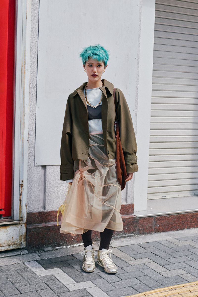 Woman with teal hair wearing an olive jacket, sheer skirt, and platform sneakers stands against a white wall in an urban setting.
