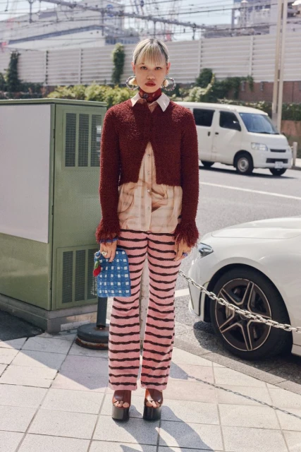 Full shot of a woman in a fashionable red and beige outfit with striped pants and platform shoes, standing on a sidewalk in a city.