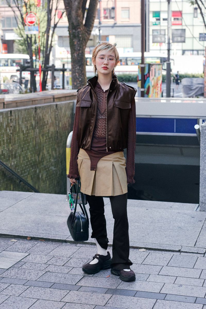A young woman with short blonde hair stands on a city sidewalk in a brown vest, top, and black leggings holding a black purse.