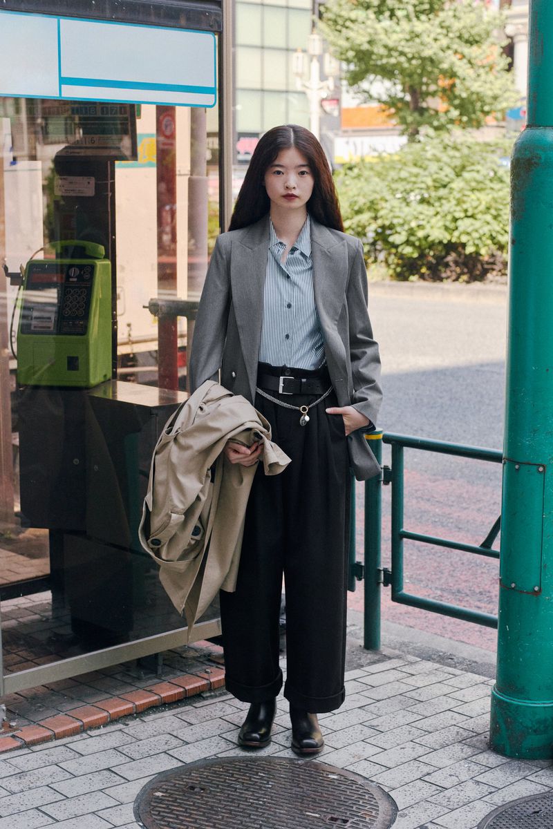 An Asian woman stands on a sidewalk in an urban area, wearing a grey blazer, light blue shirt, and black pants, holding a trench coat.