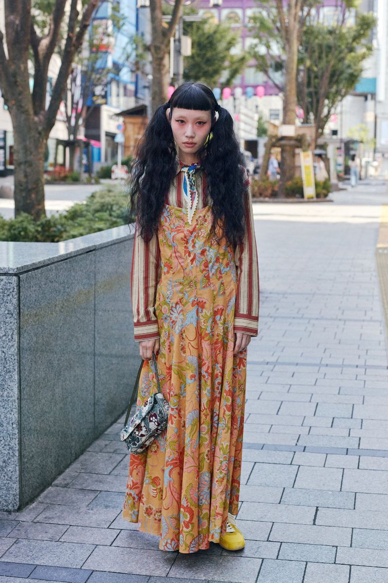 A long shot captures a young woman with pigtails, an orange floral dress, and a purse, standing on a city sidewalk with trees and buildings.