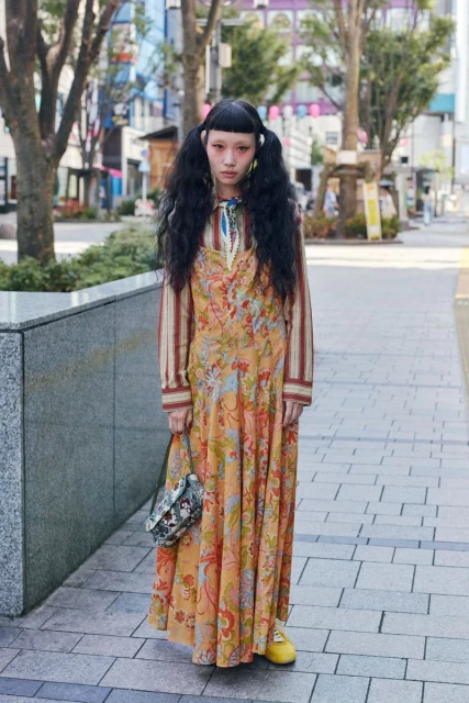 A long shot captures a young woman with pigtails, an orange floral dress, and a purse, standing on a city sidewalk with trees and buildings.