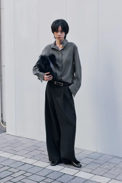 A person with short dark hair stands in front of a concrete wall in an ash-grey button-down shirt, black pants, and shiny black shoes.
