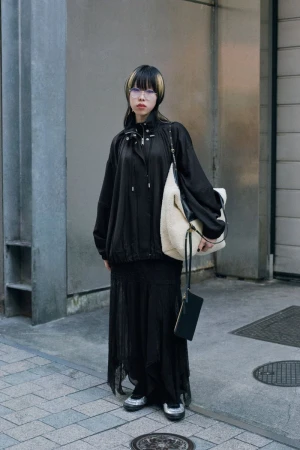 Model in all-black streetwear holding a large cream shearling shoulder bag on a city street.