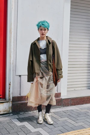 Person with vibrant teal hair wearing an olive green jacket and layered sheer skirt standing on an urban sidewalk.