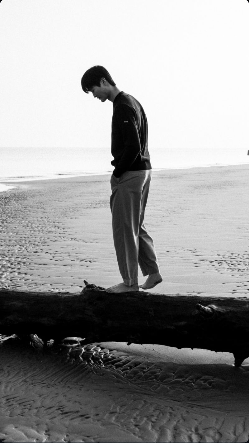 Monochrome image of a young adult man walking barefoot on a log at a beach with waves in the background, in an outdoor environment.