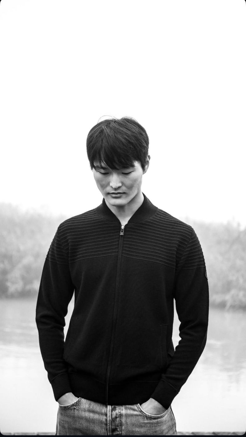 Model Cheon Jeon wearing a black zip-up knit jacket and jeans with hands in pockets outdoors in black and white.