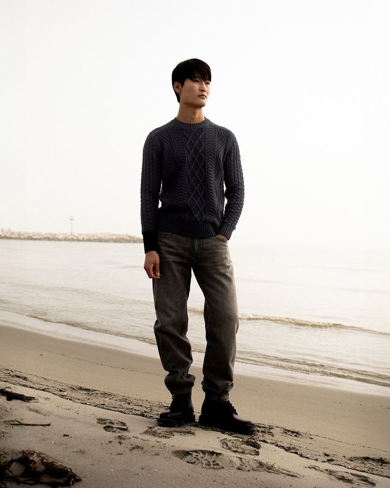 Asian man standing on a sandy beach wearing a blue sweater, jeans, and boots, looking toward the horizon on an overcast day.