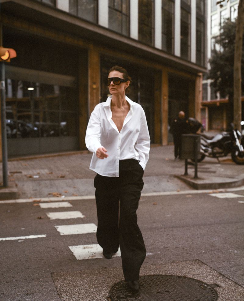 Model Tanya Ruban wearing an unbuttoned white shirt, wide-leg black trousers, and sunglasses while walking across a city street crosswalk.