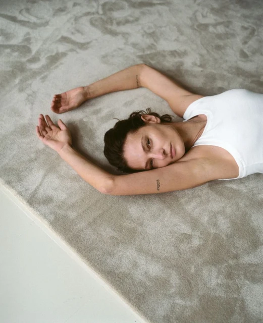 Woman lying on a textured carpet, wearing a white shirt with arms outstretched, looking at camera in an indoor environment.