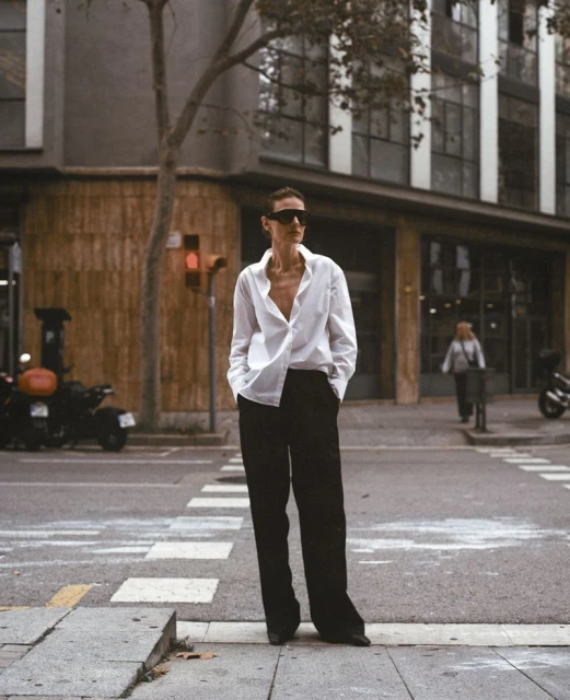 A fashionable woman in a white collared shirt and black pants confidently crosses an urban street, exuding style and modern sophistication.