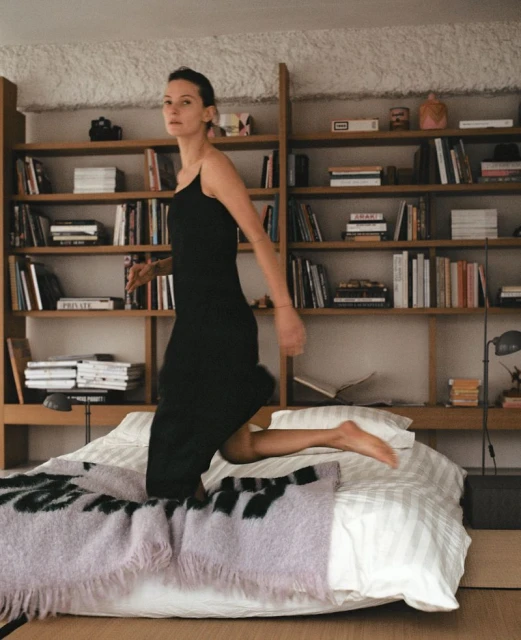 Young woman in a black dress jumps on a bed in an apartment bedroom with a bookshelf behind her, styled with a lilac blanket.