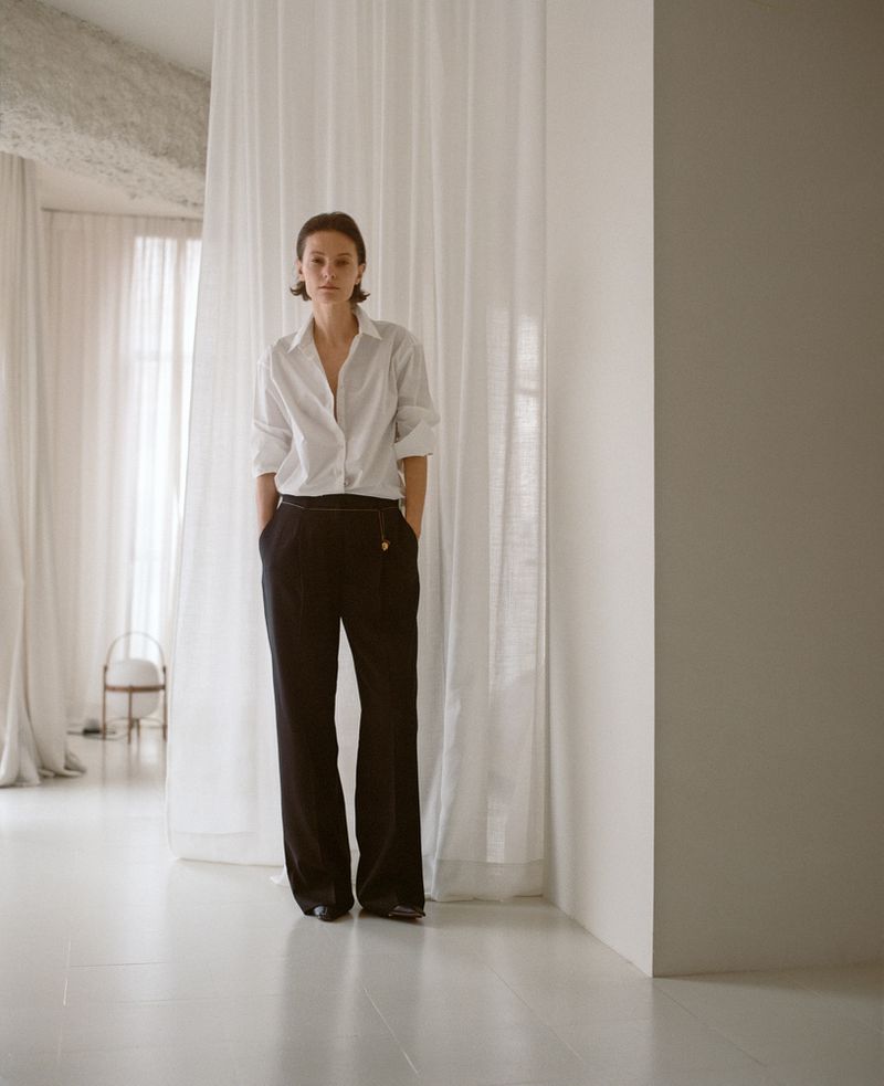 Fashionable woman in white shirt and black pants standing in a minimalist studio with sheer curtains, creating a clean, elegant aesthetic.