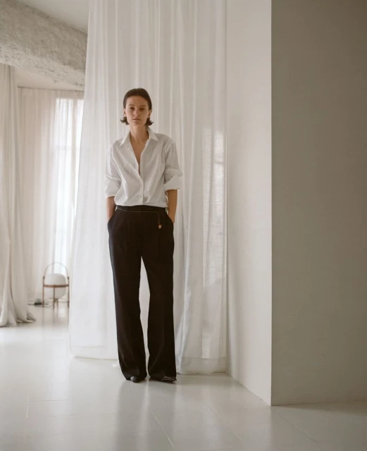 Fashionable woman in white shirt and black pants standing in a minimalist studio with sheer curtains, creating a clean, elegant aesthetic.