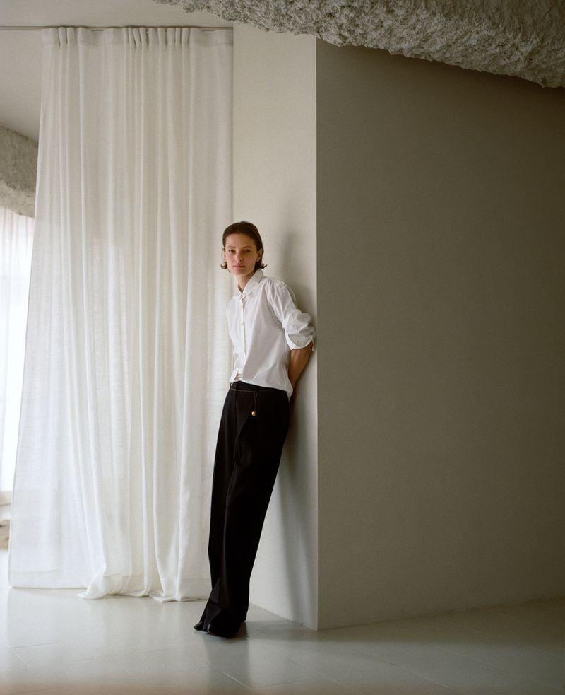 Adult leaning against a corner in a studio. She wears a white button-down shirt and black pants against a neutral background with white curtains.