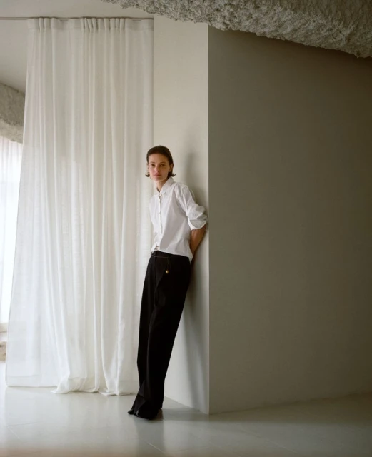 Adult leaning against a corner in a studio. She wears a white button-down shirt and black pants against a neutral background with white curtains.