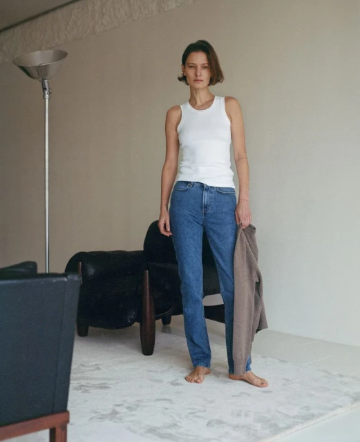A full shot of a woman standing in a room. She is wearing jeans and a white tank top, and holding a beige sweater in her hand.