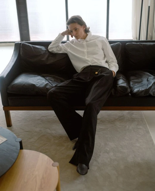 A stylish adult poses on a black leather sofa in a bright interior wearing a white button-down shirt and oversized black trousers.