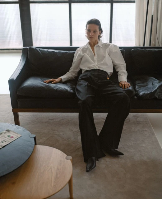 Indoor fashion portrait of a woman in a white blouse and black pants sitting on a black leather sofa with a side table.