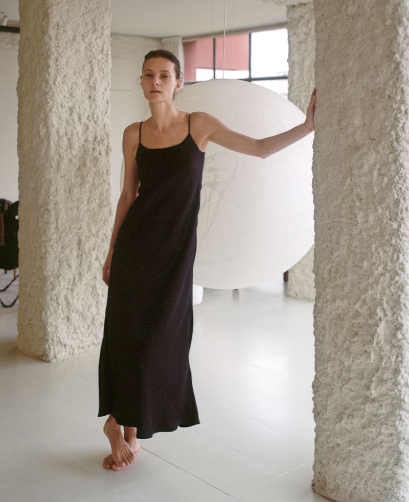 A slender woman with dark hair poses in a dark spaghetti-strap dress. She stands next to textured columns in a minimalist space.